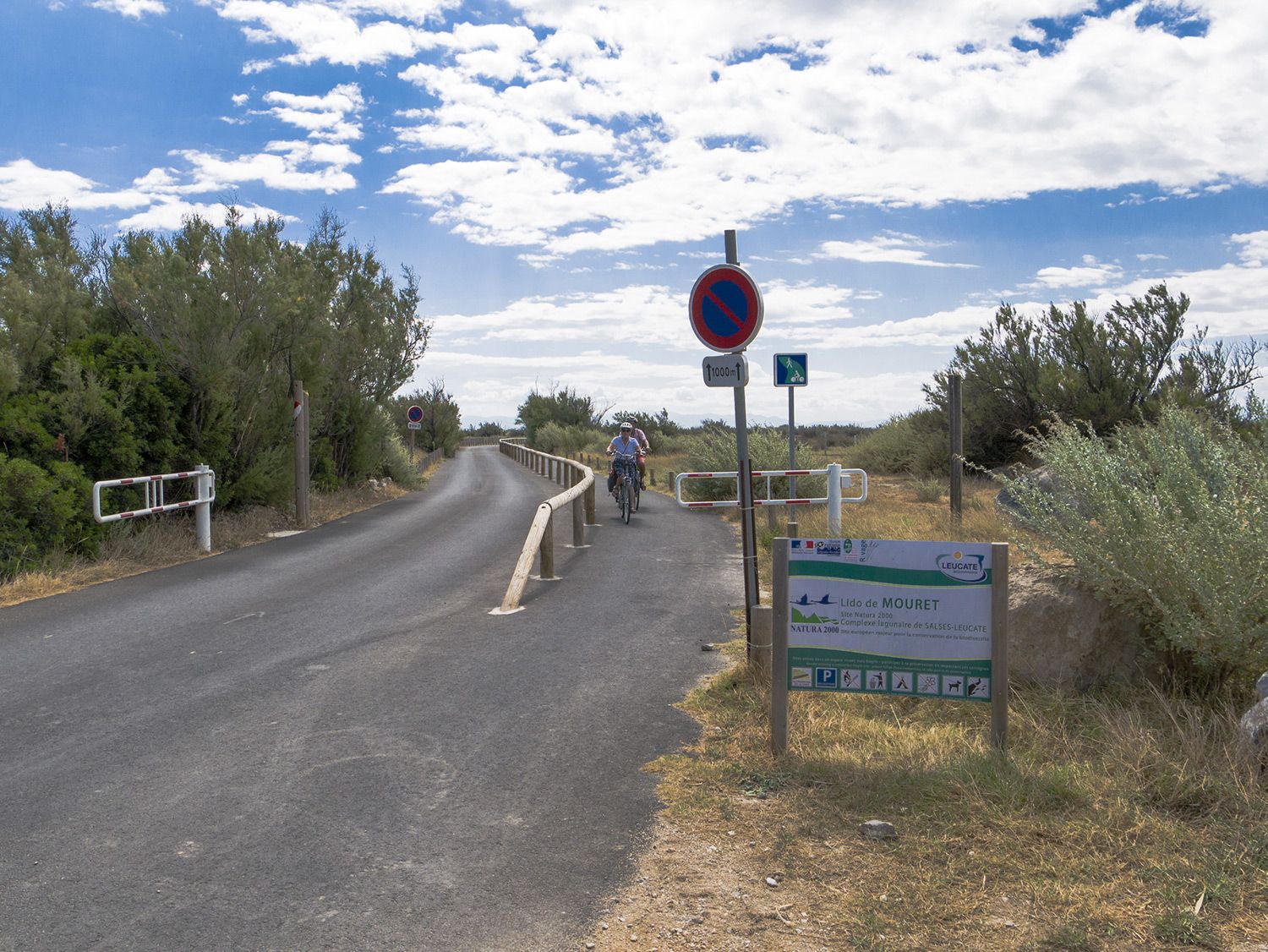 Photo d'une piste cyclable double sens sur la droite d'une route, deux personnes sont à vélo. Au premier plan un panneau indiquant le Lido de Mouret